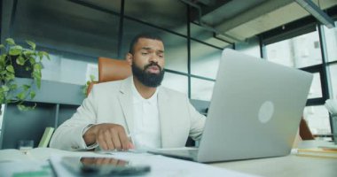 Yetenekli bir işadamı, şık bir çalışma alanında dizüstü bilgisayarıyla uğraşırken çeşitli belgeleri dikkatlice analiz eder.