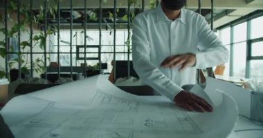 Bir mimar, çağdaş, bitki dolu ofis ortamında bina planlarını titizlikle inceler..