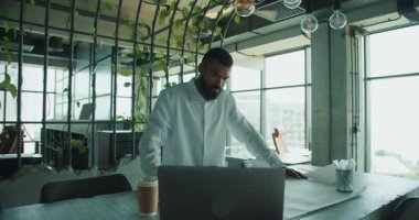 Kendini işine adamış beyaz gömlekli bir adam bitkilerle dolu modern bir ofiste dizüstü bilgisayar üzerinde çalışıyor.