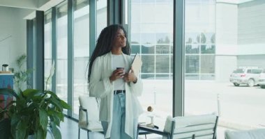 Kendine güvenen profesyonel bir kadın, elinde bir tablet ve bir fincan kahveyle modern ve şık bir ofis ortamında duruyor.