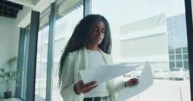 Bir kadın, parlak ve çağdaş ofis ortamında çeşitli kağıtları dikkatle ve dikkatle gözden geçirir.