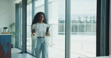 Profesyonel bir kadın, parlak bir ofiste dururken dizüstü bilgisayar ve kahve fincanı tutuyor.