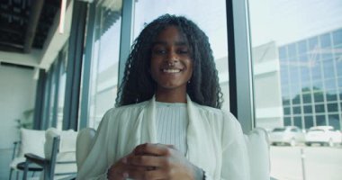 Kıvırcık saçlı genç bir kadın modern bir ofiste neşeli bir şekilde gülümsüyor, samimi ve davetkar bir ortam yaratıyor.