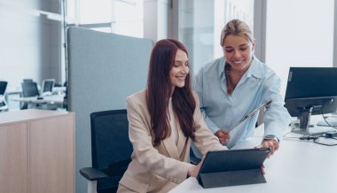 İki profesyonel kadın, modern bir çalışma ortamında tablet kullanırken üretken bir tartışma içindeler.