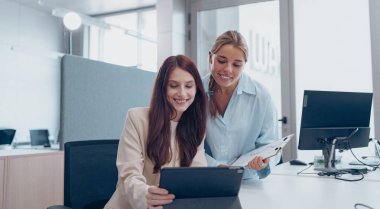 İki kadın bir tablete bakıyor, şık bir ofis ortamında işbirliği yapıyorlar, takım çalışması ve profesyonellik sergiliyorlar..