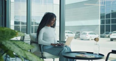 Odaklanmış ve kararlı bir kadın, modern ve şık bir ofis odasında dizüstü bilgisayarla otururken işine dalmış durumda.