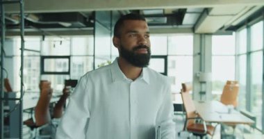 Kendine güvenen beyaz gömlekli bir adam modern ve şık bir ofis alanında yürürken görülüyor.