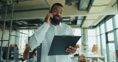 Beyaz gömlekli gülümseyen bir adam, çağdaş bir ofiste belgeleri incelerken bir telefon görüşmesi yapıyor..