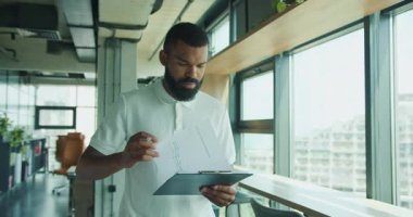 Kendini işine adamış ve odaklanmış bir adam şık, modern bir ofis ortamında otururken tablet ve kağıt kullanıyor.