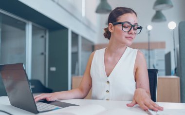 Şık ve odaklanmış bir kadın, parlak ve çağdaş bir ofis içinde özenle dizüstü bilgisayarının başında çalışıyor.