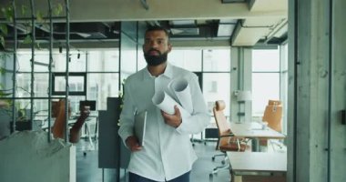 Kendini işine adamış ve işine odaklanmış bir profesyonel elinde parlak, modern bir ofis alanında planlar ve bir dizüstü bilgisayar tutuyor.