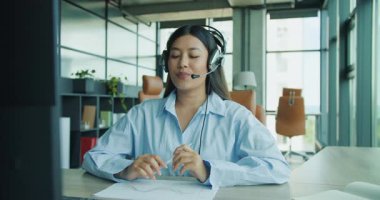 Kulaklık takan genç bir profesyonel aktif olarak meşgul ve parlak bir ofis alanında bir video görüşmesine katılıyor. Yüksek kalite 4k görüntü