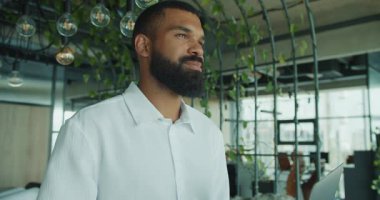 Kendine güvenen bir işadamı canlı bir ofiste gururla duruyor. Yeşillik ve modern profesyonelliği gözler önüne seriyor.