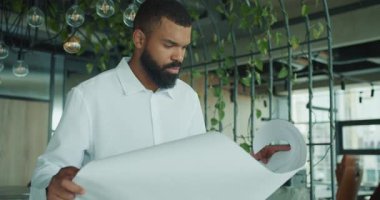 Genç bir adam, mimari planları bitkilerle dolu parlak bir çalışma alanında gözden geçirir, yaratıcılığı gözler önüne serer.