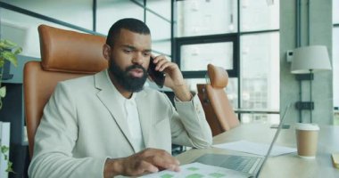 Keskin giyimli takım elbiseli bir iş adamı, modern bir ofiste dizüstü bilgisayarı üzerinde çalışırken bir telefon görüşmesi yapar.