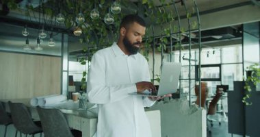 Kendini işine adamış bir profesyonel şık, bitkisel bir ofis ortamında otururken dizüstü bilgisayar kullanıyor.