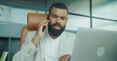 Takım elbiseli kararlı bir adam, modern bir ofisteki bir telefon görüşmesinde bilgisayarını inceliyor.