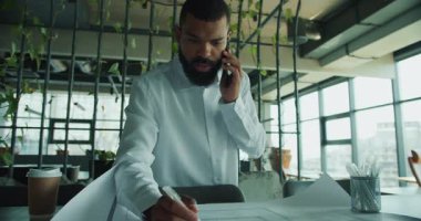 Çağdaş ofis ortamında bir işadamı, belgeleri incelerken telefonda planları tartışıyor..