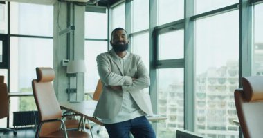 Kendine güvenen profesyonel bir adam, parlak bir ofiste gülümsüyor. Geniş pencereli, ışık saçan bir tarzı ve kendine güveni var.