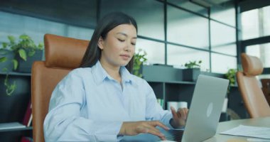 Şık bir ofis ortamında özenle dizüstü bilgisayara yazı yazan odaklanmış genç bir kadın canlı yeşillikle süslenmiş. Yüksek kalite 4k görüntü