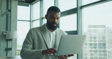 Şık takım elbiseli odaklanmış bir adam bilgisayarını parlak ve çağdaş bir ofis alanında etkin bir şekilde kullanıyor.