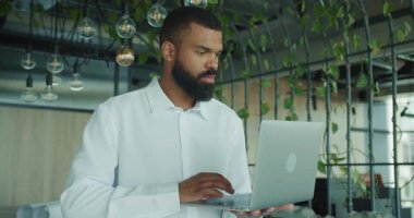 Genç bir profesyonel, yeşillik ve bitkilerle çevrili şık bir çalışma alanında dizüstü bilgisayarına odaklanıyor.