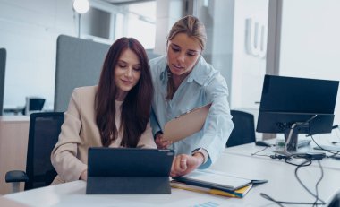 Modern bir ofis ortamında, iki kadın tablet kullanırken fikirleri tartışır.