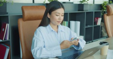 Profesyonel bir kadın, ofisinde dizüstü bilgisayarı olan bir masada otururken telefonunu kullanıyor. Yüksek kalite 4k görüntü