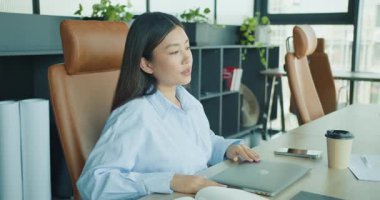 Mavi gömlekli odaklanmış genç bir kadın, şık ve modern bir ofis alanında özenle dizüstü bilgisayarıyla çalışıyor. Yüksek kalite 4k görüntü