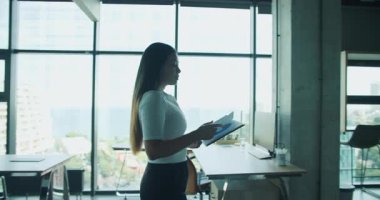 Profesyonel bir kadın ofiste özgüvenle durur, elinde defteriyle pencereden dışarı dikkatlice bakar. Yüksek kalite 4k görüntü