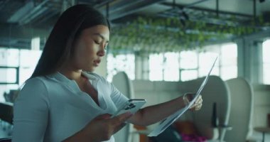 Kendini işine adamış profesyonel bir kadın telefonunu modern bir çalışma alanında kullanırken önemli evrakları gözden geçirir.