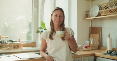 Bir kadın, güzel doğal ışıkla dolu büyüleyici ve rahat bir mutfakta sıcak kahvesinin tadını çıkarıyor.
