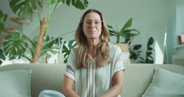 Kendine güvenen, gülümseyen bir kadın yeşil ve modern dekorlarla çevrili şık bir oturma odasında oturur ve sıcaklık yaratır.