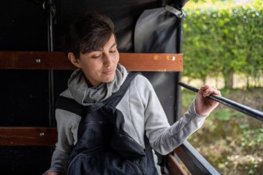 Perulu bir kadın eşofman üstü giyiyor ve sırt çantasını önünde taşıyor. Arka planı ve kırsal kesimde olan bir arabanın arkasında.