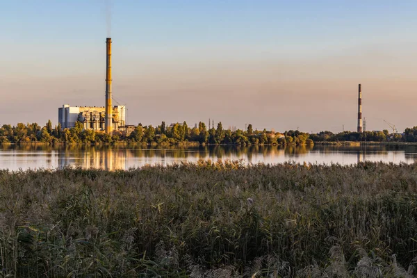 The beauty of a lake at sunset is marred by the presence of an old garbage incineration plant. Its smoking smokestacks emit air pollution, including from the energy generated from waste.