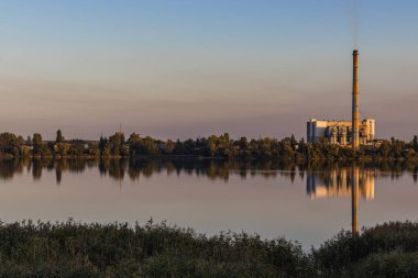 Bir göl kıyısındaki atık işleme tesisi atıklardan enerji üretir, fakat atıklara eşlik eden bacalar atmosfere hava kirliliği salar. Renkli gün batımı gökyüzü