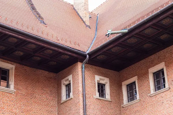 Krakow 'daki Collegium Maius' un mimari detayları. Kırmızı fayanslı çatı, süslü ahşap tavan ve dekoratif bir heykel. Tuğla duvar ve pencereler binaların tasarımını ön plana çıkarıyor