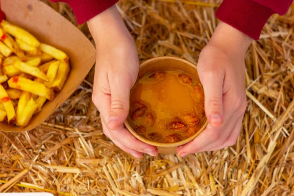 hands hold a cup of pumpkin soup next to french fries (chips) in a cardboard plate on a table with hay. traditional country food at a farmer's festival.