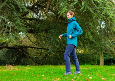 Spor ceketli genç bir kadın ormanın yakınındaki bir çayırda yürüyor. Doğa, yürüyüş, aktif yaşam tarzı, kamp.