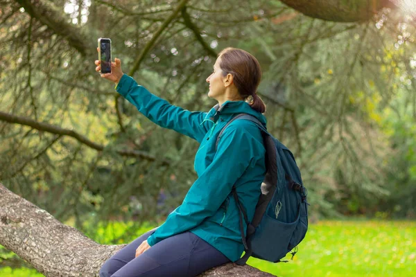 a young woman in warm clothes sits on a tree branch in the forest and takes a selfie on her mobile phone. nature, hiking, active lifestyle, camping