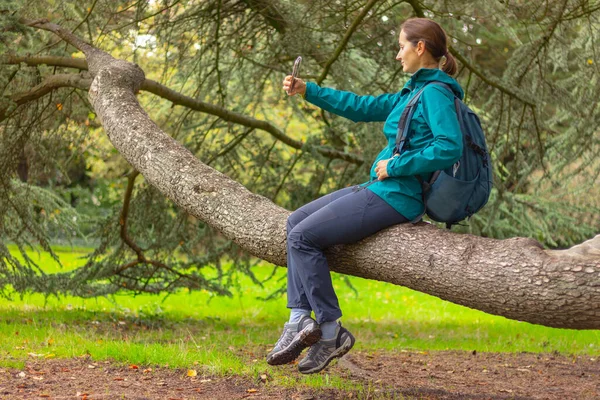 young woman with mobile phone in sport clothes and backpack sits on a tree branch in the forest and takes a selfie. nature, hiking, active lifestyle, camping