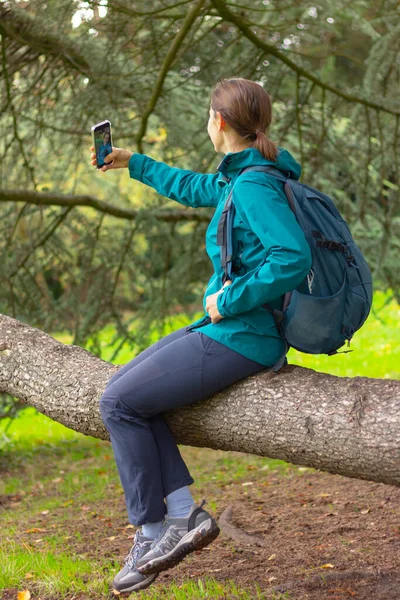 young woman with mobile phone in sport jacket sits on a tree branch in the forest and takes a selfie. nature, hiking, active lifestyle, camping