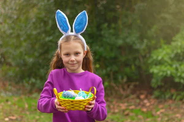 A cute girl with rabbit ears and basket of colored eggs on natural background. Easter bunny fun costume, childhood