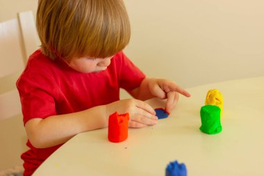 Açık renk saçlı, kırmızı tişörtlü bir çocuk masada plasticinle oynuyor. çocuk eğlencesi, eğlence, aktiviteler, eğitim oyunları.