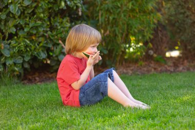 Çocuk parktaki çimlerin üzerinde oturmuş karpuz yiyor.