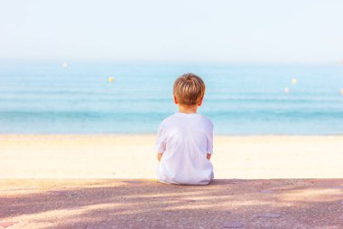 Bir çocuk deniz kıyısında oturuyor, arka manzaralı.