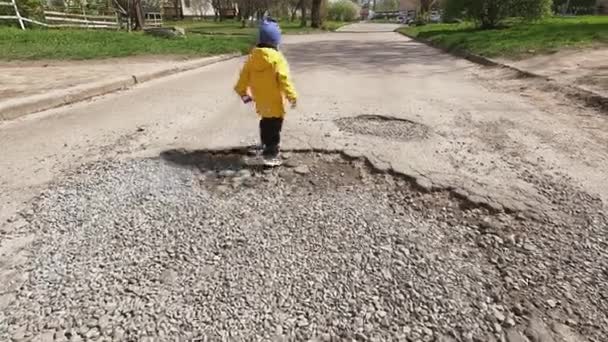 子供は大きなピットとアスファルトの道路に沿って歩く コピースペース 道路の修復 都市インフラ 子供の交通安全 途中の障害物の克服 — ストック動画