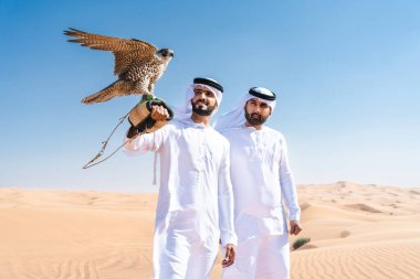 Geleneksel emirati Arap kandurası giyen iki orta doğulu adam çölde kaynaşıyor ve bir şahin kuşu tutuyor - Arap Müslüman arkadaşlar Dubai 'de kum tepelerinde buluşuyorlar.