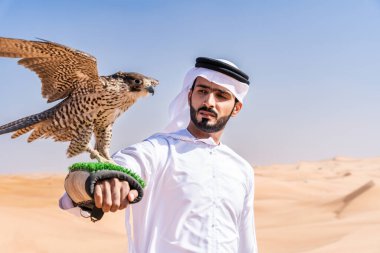 Ortadoğulu bir adam çölde geleneksel emirati Arap kandurası giyiyor ve Dubai 'deki kum tepelerinde Arap Müslüman bir şahin kuşu tutuyor.