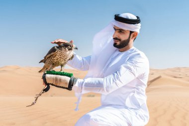 Ortadoğulu bir adam çölde geleneksel emirati Arap kandurası giyiyor ve Dubai 'deki kum tepelerinde Arap Müslüman bir şahin kuşu tutuyor.